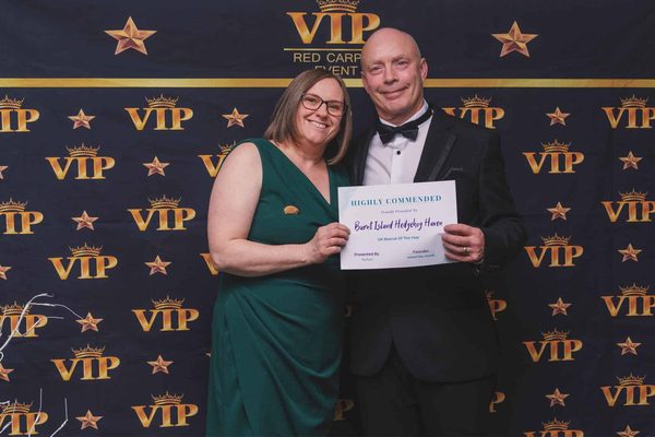 A woman in a green dress and a man in a dinner suit hold a Highly Commended certificate in front of a VIP backdrop.