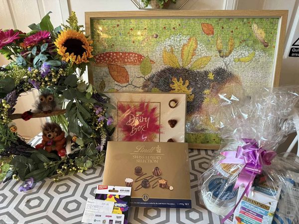 Raffle prizes on a table: a flower wreath with felt animals, a hedgehog jigsaw, chocolates, a voucher and a wrapped gift basket.
