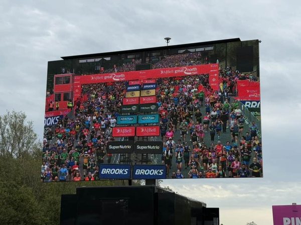 Large outdoor screen showing a crowd of runners at a race start, with sponsor banners for Brooks and SuperNutrio.