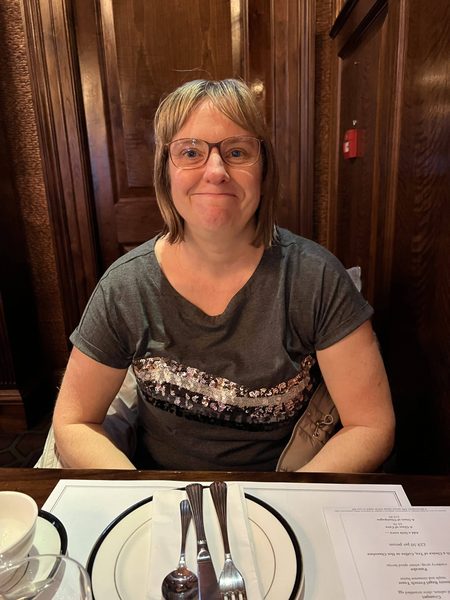 Sharon, in a grey sparkly top, sits at a restaurant table set with cutlery and a plate, with wood-panelled walls behind.
