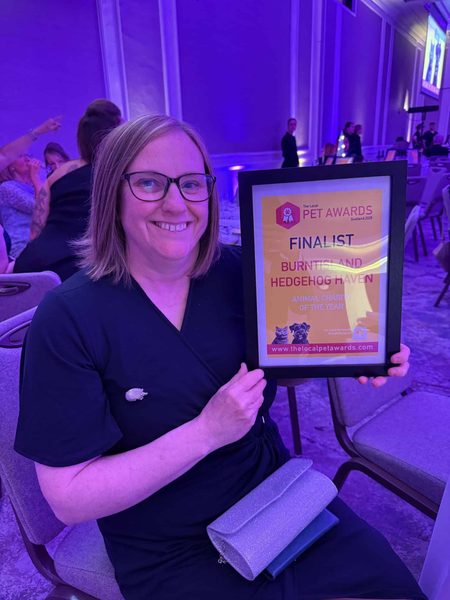 Smiling woman in glasses sits holding a framed Finalist certificate for Burntisland Hedgehog Haven at an event with purple lighting.