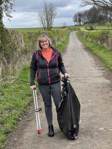 Sharon stands on a country lane holding a litter picker and a full black bin bag, smiling.