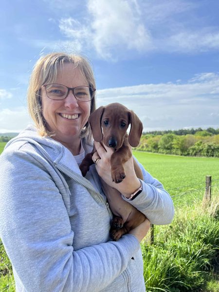 Sharon smiles in a grey hoodie, holding a dachshund puppy in front of green fields and a blue sky.