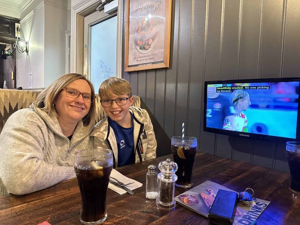 A smiling woman and a boy in glasses sit at a pub table with two glasses of cola, with a football match on the TV behind.