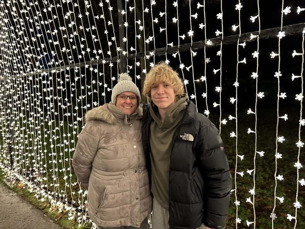 A woman in a bobble hat and a young man in a black puffer jacket stand in front of a curtain of white fairy lights at night.
