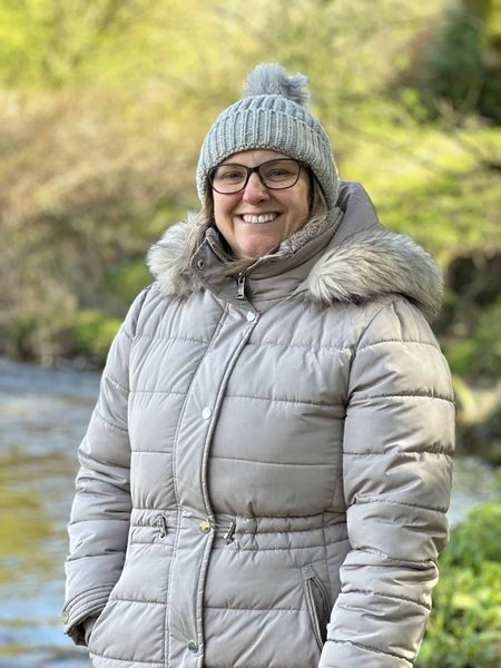 Smiling woman in a grey bobble hat, glasses and padded coat stands by a stream with green foliage behind.