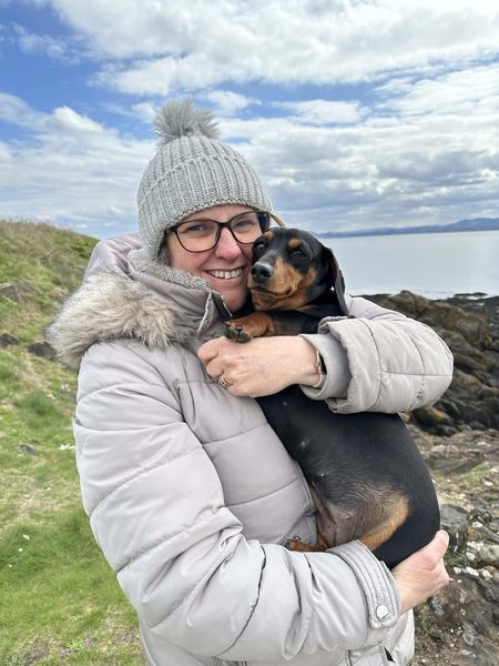 Smiling woman in a bobble hat, glasses and padded coat hugs a black and tan dachshund by the sea.