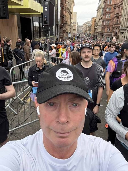 Selfie of a man in a Burntisland Hedgehog Haven cap in a busy street full of runners queuing behind barriers.