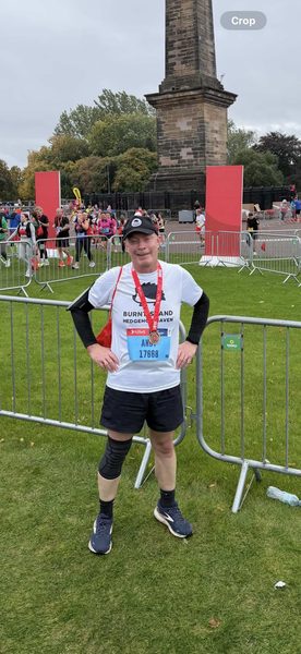 A man in a Burntisland Hedgehog Haven T-shirt, cap and race medal stands by barriers near a stone monument.