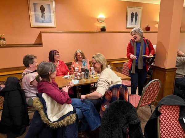 Women sit around a pub table laughing and chatting while another woman in a red jacket stands holding papers.