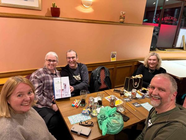 Five smiling people around a pub table with drinks, bingo pens and a gift bag, one holding up a boxed prize.