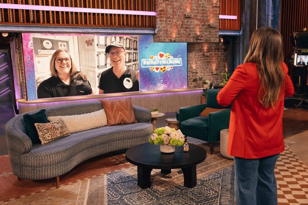 Woman in a red jacket faces a studio screen showing two smiling people in black tops, on a set with a sofa and coffee table.