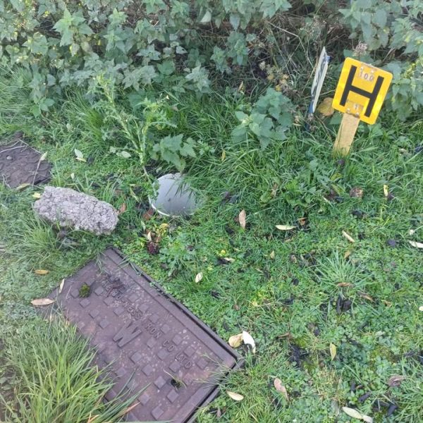 Uncovered round pipe in grass between two metal covers, with a yellow hydrant marker post and a piece of rubble nearby.