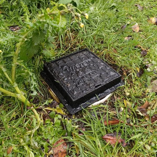 Square black metal cover sitting over a hole in long grass and nettles.