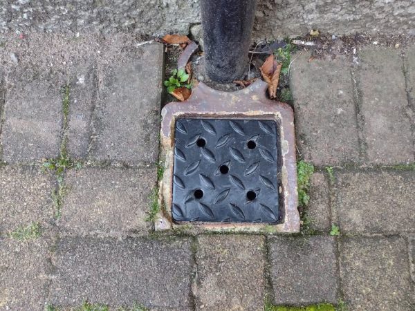 Black metal drain cover with small round holes beside a downpipe, set in grey paving.